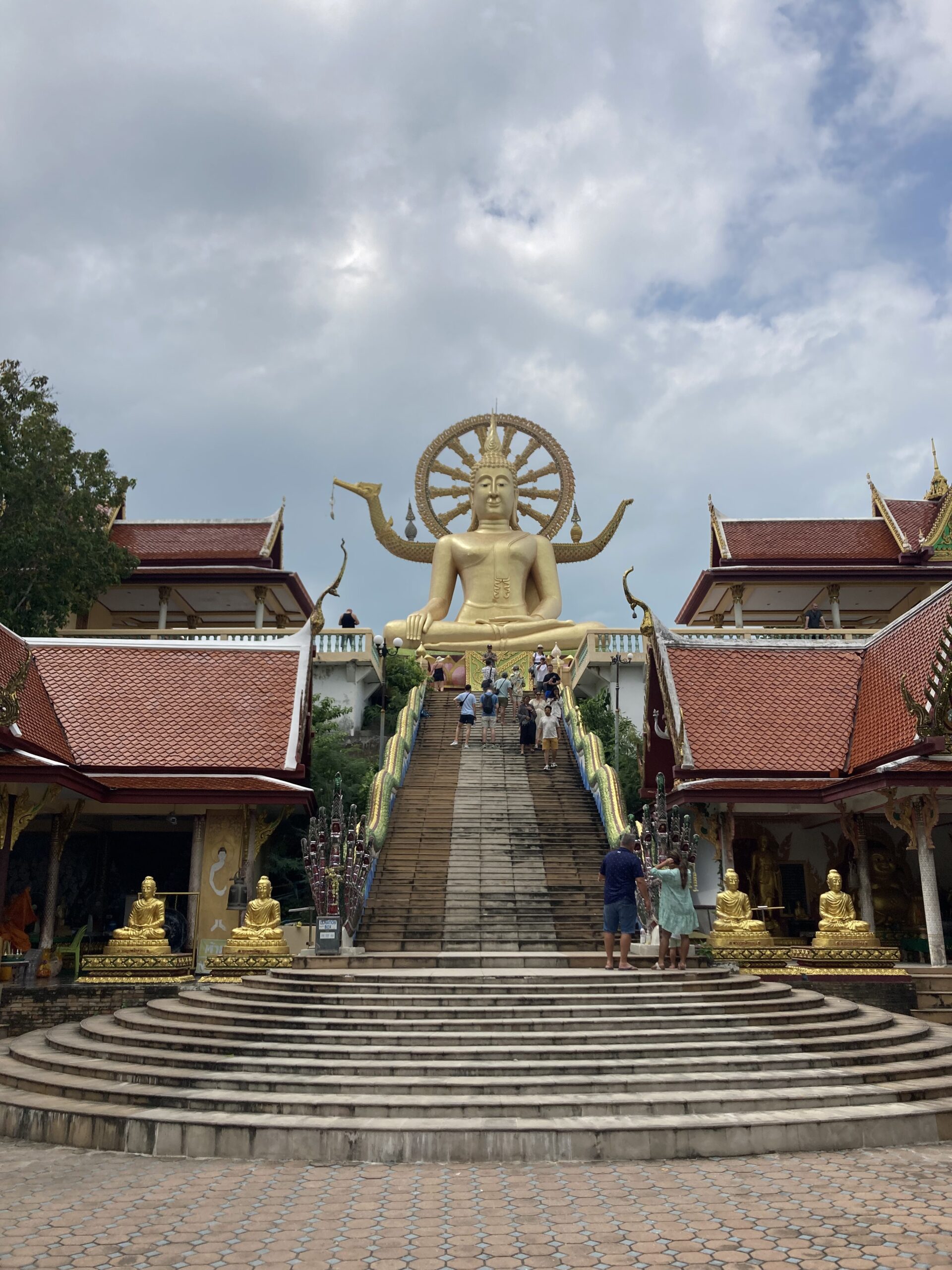 Eine große goldene Buddhastatue steht am oberen Ende einer breiten Treppe, flankiert von kunstvollen Gebäuden mit roten Dächern. Mehrere Menschen, die vielleicht Geschichten von ihrer letzten Segelreise nach Griechenland erzählen, steigen die Treppe hinauf. Der Himmel darüber ist bewölkt mit blauen Flecken.