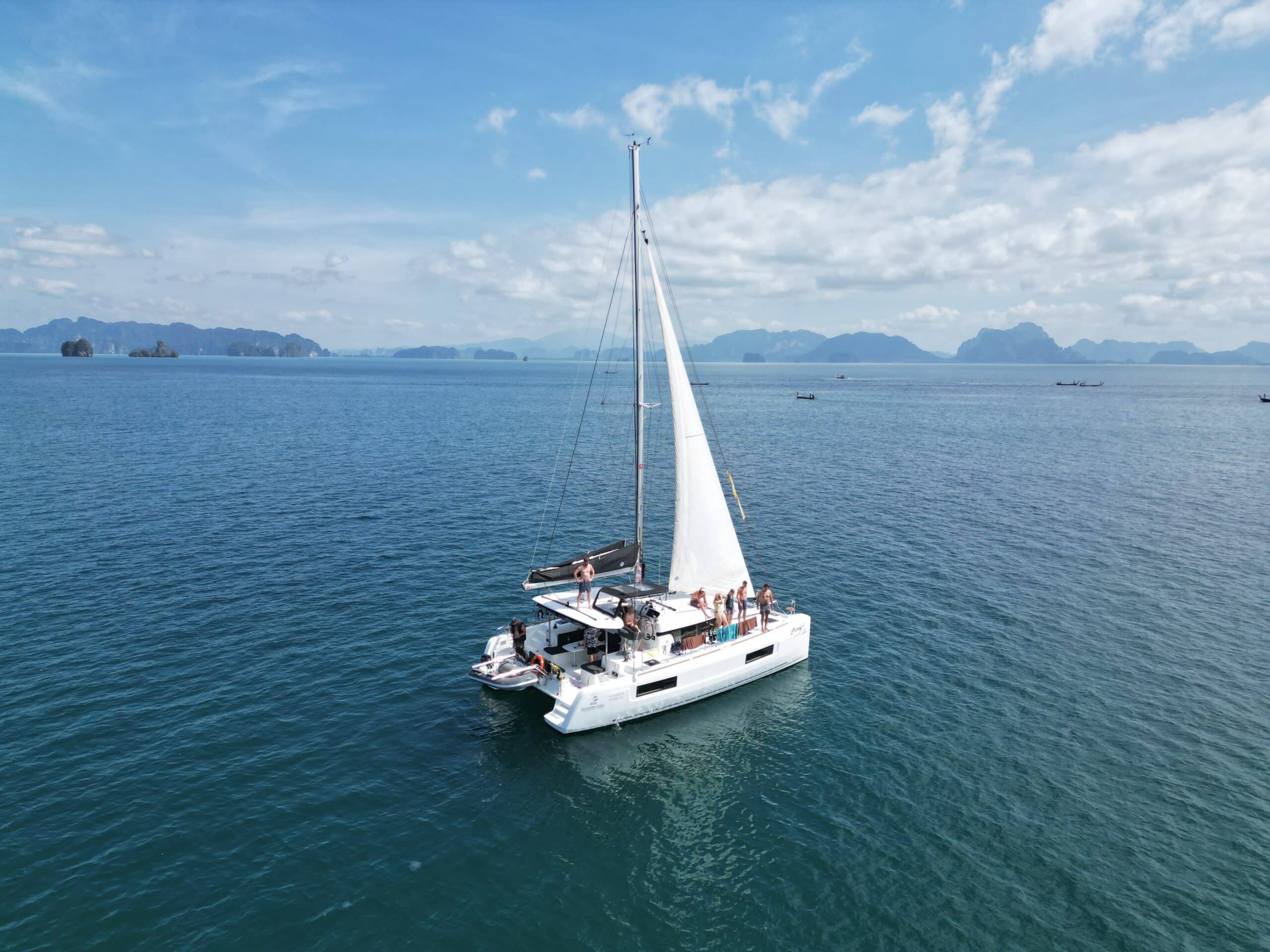 Ein weißes Segelboot mit einer Gruppe von Menschen an Bord kreuzt in den ruhigen blauen Gewässern vor Phuket, Thailand, mit einer Kulisse aus fernen Inseln unter einem teilweise bewölkten Himmel. Das Segel ist vollständig ausgefahren und die Szene zeigt ein ruhiges und malerisches Segeln.