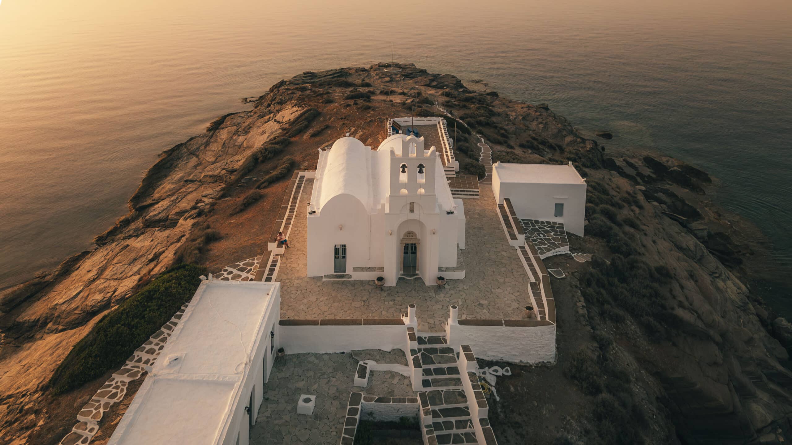 Luftaufnahme einer weißen Kapelle mit Bogenfenstern und einem kleinen Glockenturm nahe der Klippenkante mit Blick auf ein ruhiges Meer bei Sonnenuntergang. Die felsige Landschaft und die Steinpfade rund um die Kapelle tragen zur friedlichen, malerischen Atmosphäre bei – perfekt für alle, die eine Segelreise nach Griechenland unternehmen.