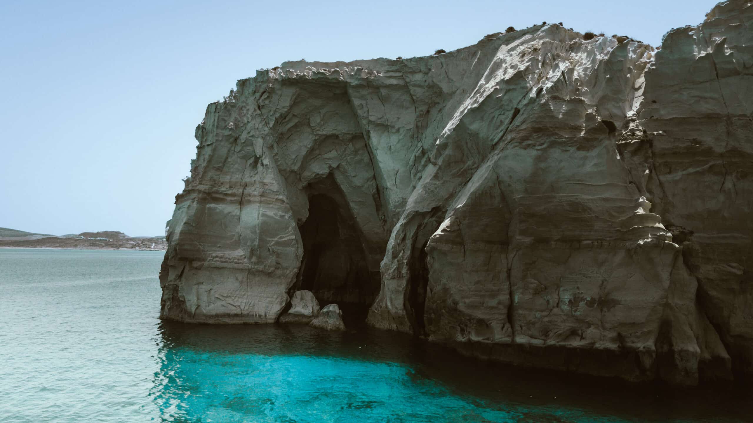 Eine große Felsklippe mit einem Höhleneingang liegt am türkisfarbenen Meer, ein malerischer Anblick, den man oft während einer Segelreise nach Griechenland genießt. Die zerklüftete Oberfläche der Klippe wirft Schatten und man kann verschiedene Vögel auf der Spitze sehen. Darüber erstreckt sich der klare blaue Himmel.