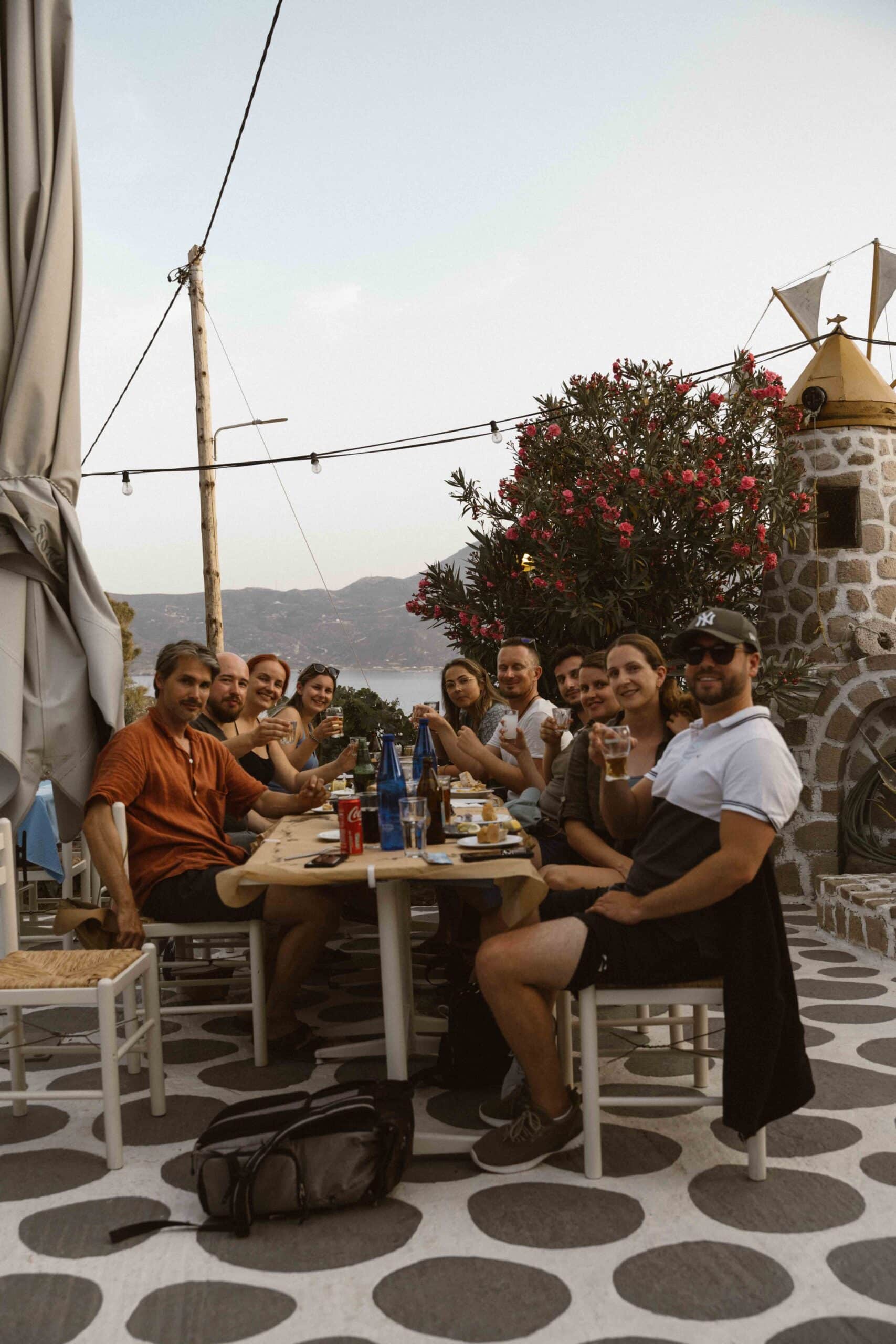 Eine Gruppe von Menschen sitzt um einen langen Tisch im Freien, lächelt und stößt mit den Gläsern an. Die Kulisse besteht aus einem Steinboden, einer Windmühle, blühenden Büschen und Lichterketten über dem Kopf. Im Hintergrund sind sanfte Hügel unter einem klaren Himmel zu sehen, was an eine ruhige Segelreise nach Griechenland erinnert.