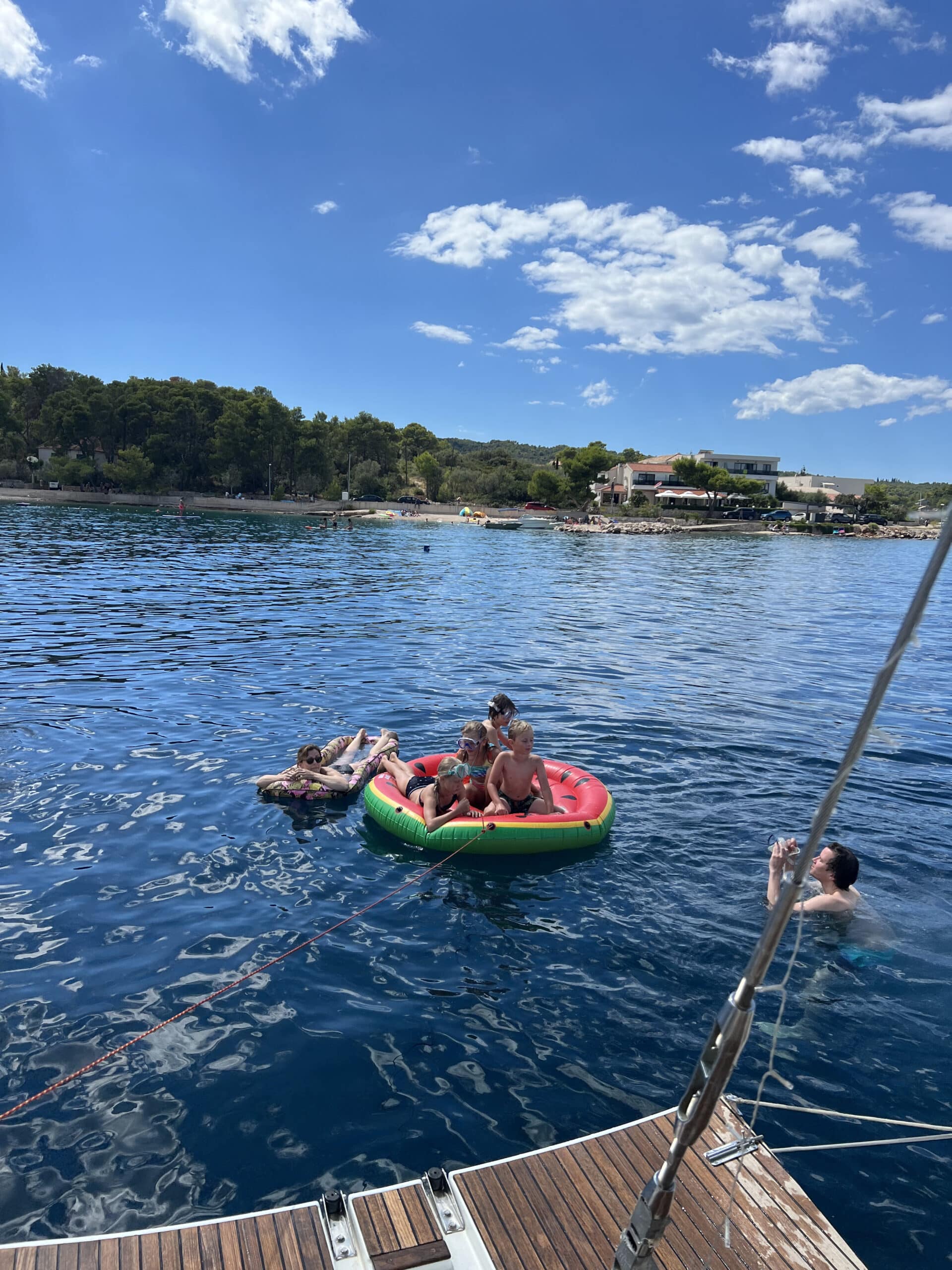 Vier Menschen genießen einen sonnigen Tag auf dem Wasser. Drei von ihnen sitzen auf einem großen, aufblasbaren rot-grünen Schwimmkörper, während eine Person in der Nähe schwimmt. Im Hintergrund ist die Uferlandschaft mit Bäumen, Gebäuden und ein paar Menschen zu sehen, was den Charme des familienfreundlichen Segeltörns im Mittelmeer hervorruft.