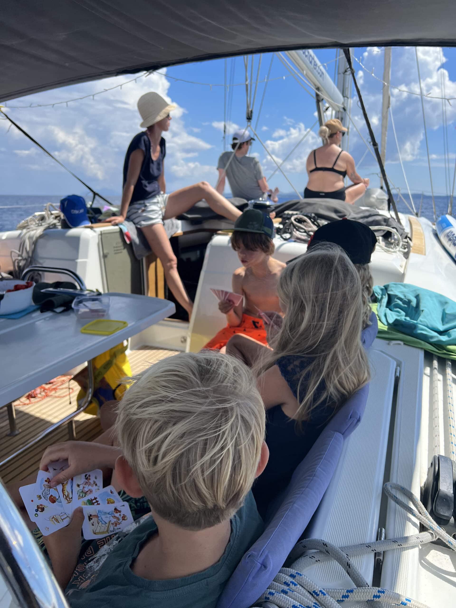 Eine Gruppe von Menschen, darunter Kinder und Erwachsene, entspannt sich auf einem Segelboot unter einem teilweise bewölkten Himmel. Die Kinder sind mit einer Aktivität beschäftigt und schauen sich Aufkleber an, während die Erwachsenen sich entspannen und unterhalten. Ausrüstung, Seile und Segelausrüstung sind sichtbar. Die Atmosphäre ist heiter während dieser familienfreundlichen Segeltörns im Mittelmeer.