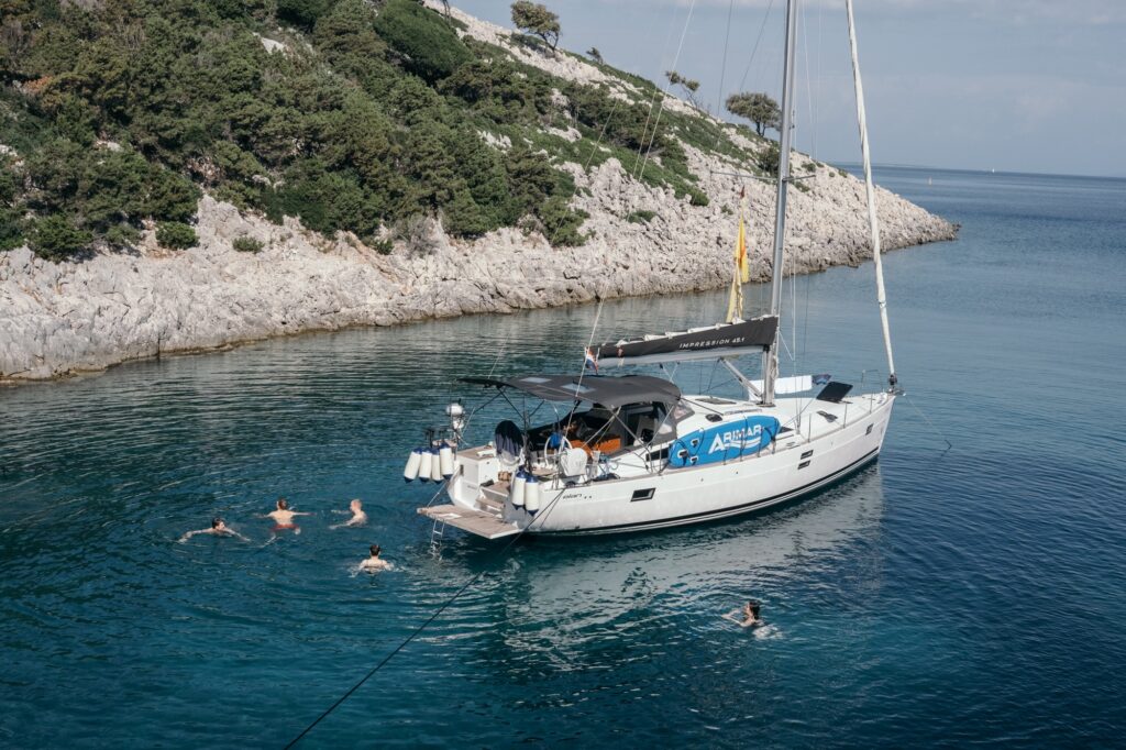 Ein weißes Segelboot mit schwarzem Verdeck liegt in einer klaren, ruhigen blauen Bucht nahe einer felsigen, von Bäumen gesäumten Küste vor Anker. Mehrere Menschen schwimmen um das Boot herum und genießen das ruhige Wasser und die natürliche Umgebung. Rundreise plus Badeurlaub: Die perfekte Kombinationsreise
