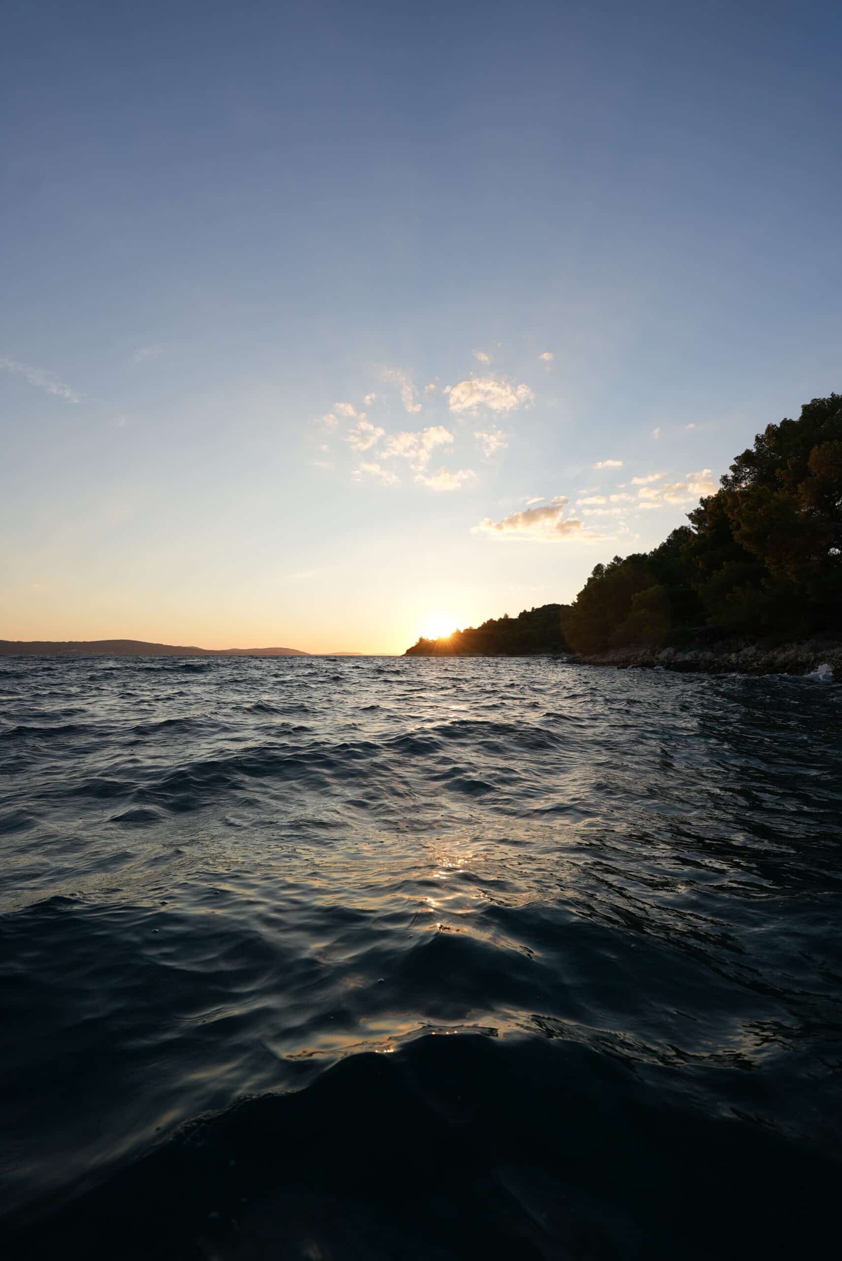 Ein ruhiger Blick auf das Meer bei Sonnenuntergang mit sanften Wellen im Vordergrund. Die Sonne steht tief am Horizont und wirft ein warmes, goldenes Licht auf das Wasser. Rechts ist eine mit Bäumen bedeckte Klippe zu sehen, die sich bis ins Meer erstreckt und an die Abenteuer von Mitsegeln Kroatien erinnert. Am Himmel sind ein paar leichte Wolken.