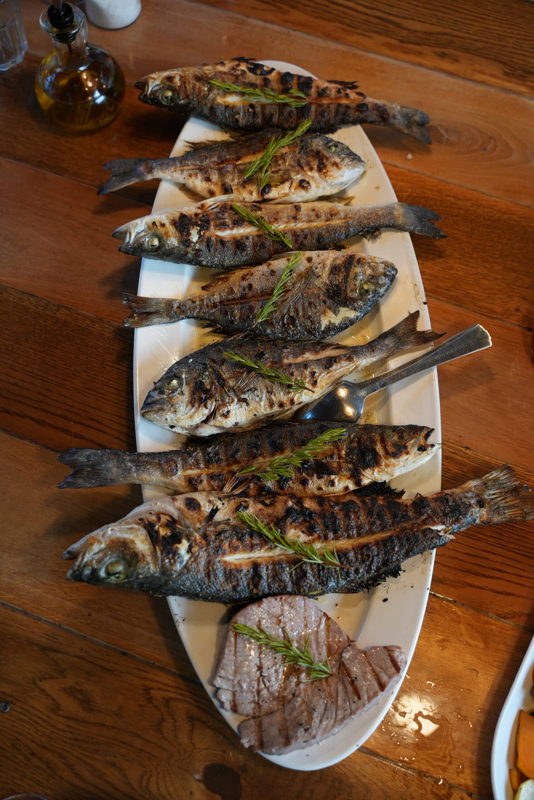 Auf einem Holztisch liegt eine weiße Platte mit sechs gegrillten ganzen Fischen, garniert mit Kräutern, die an die frischen Aromen von Mitsegeln in Kroatien erinnern. Zwischen den Fischen liegen zwei silberne Besteckteile. Auf dem Boden der Platte liegen mehrere Stücke gegrilltes Fischfilet. Im Hintergrund stehen Flaschen und Geschirr.