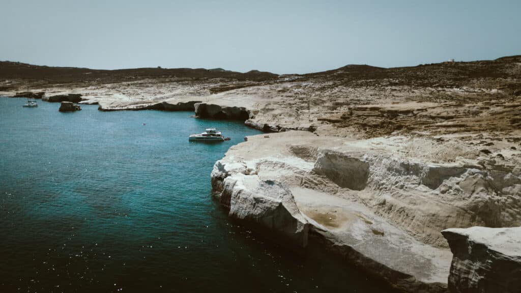 Eine ruhige Küstenszene mit dem klaren türkisfarbenen Wasser und den schroffen, weißen Felsklippen der Kykladen. Ein paar Boote schwimmen in Ufernähe, ein größeres liegt nah an den Felsen vor Anker. Die Landschaft im Hintergrund ist trocken, mit minimaler Vegetation, unter einem klaren blauen Himmel.