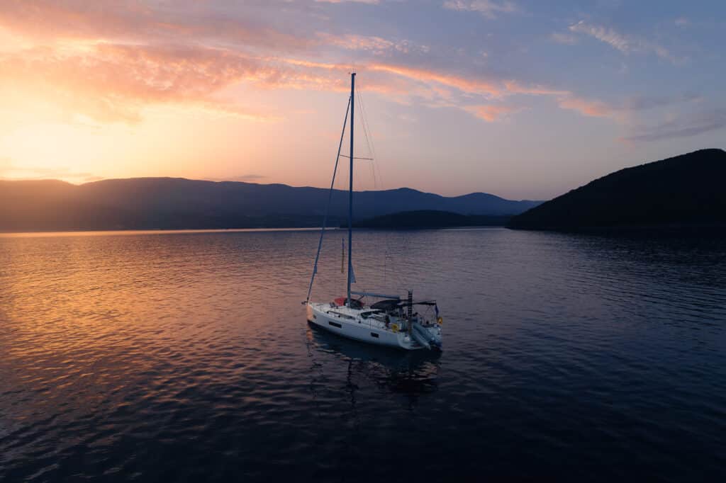 Ein Segelboot schwimmt friedlich auf ruhigem Wasser während eines leuchtenden Sonnenuntergangs und träumt vielleicht von einer Atlantiküberquerung. Der Himmel ist in Orange-, Rosa- und Lilatönen gefärbt. Im Hintergrund zeichnen sich Berge und Hügel ab, was zu der ruhigen und malerischen Szene beiträgt. Sieht nach einem perfekten, umweltfreundlichen Segelurlaub aus.