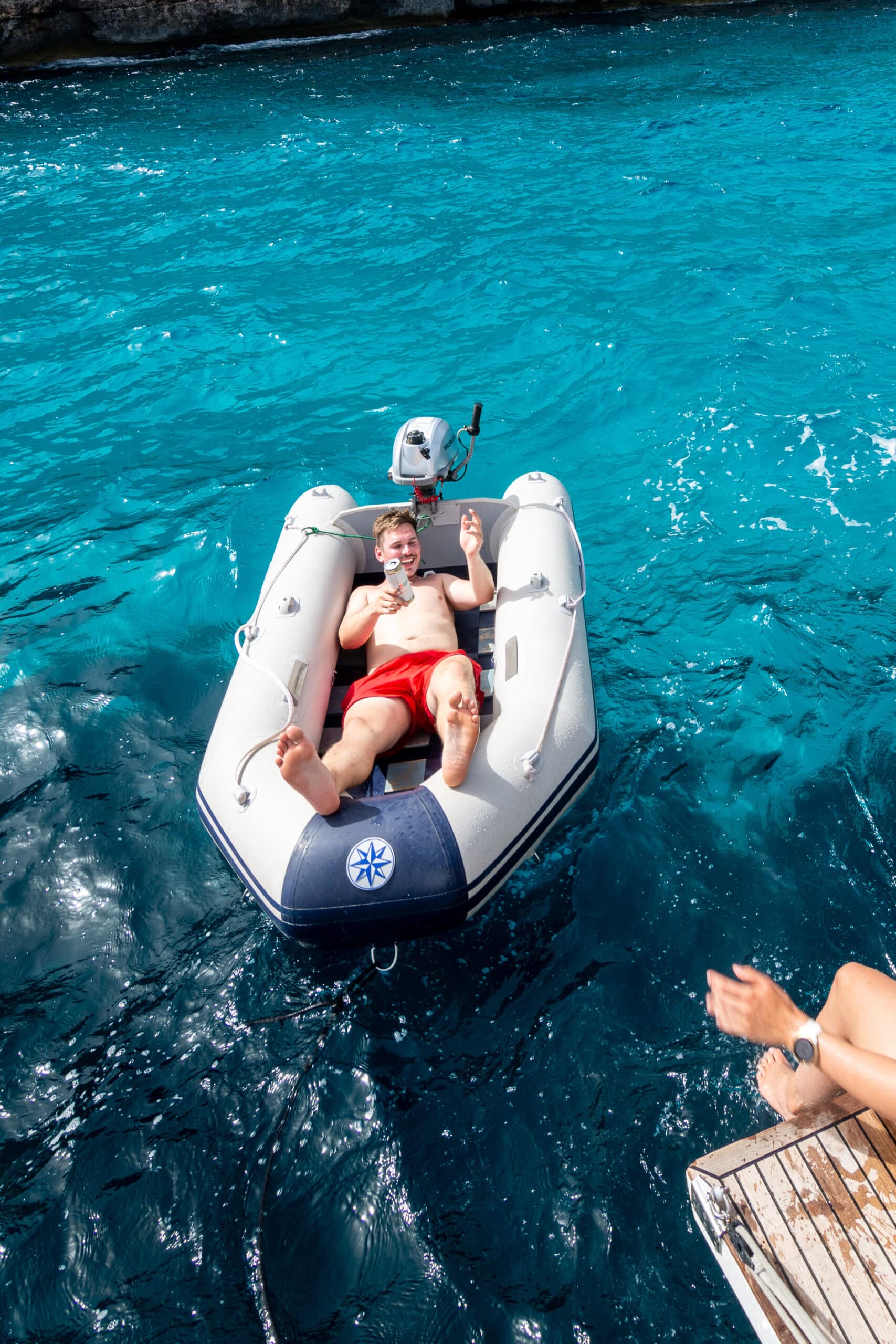 Eine Frau sitzt auf einer Bootsplattform, lässt ihre Beine in Richtung Wasser baumeln und winkt zwei lächelnden Personen in einem kleinen Schlauchboot zu. Sie treiben im hellen Sonnenlicht auf klarem, türkisfarbenem Wasser in der Nähe einer Felsklippe. Auf der Klippe wachsen Bäume und Sträucher – eine idyllische Kulisse für ihr Segeltörn-Abenteuer.