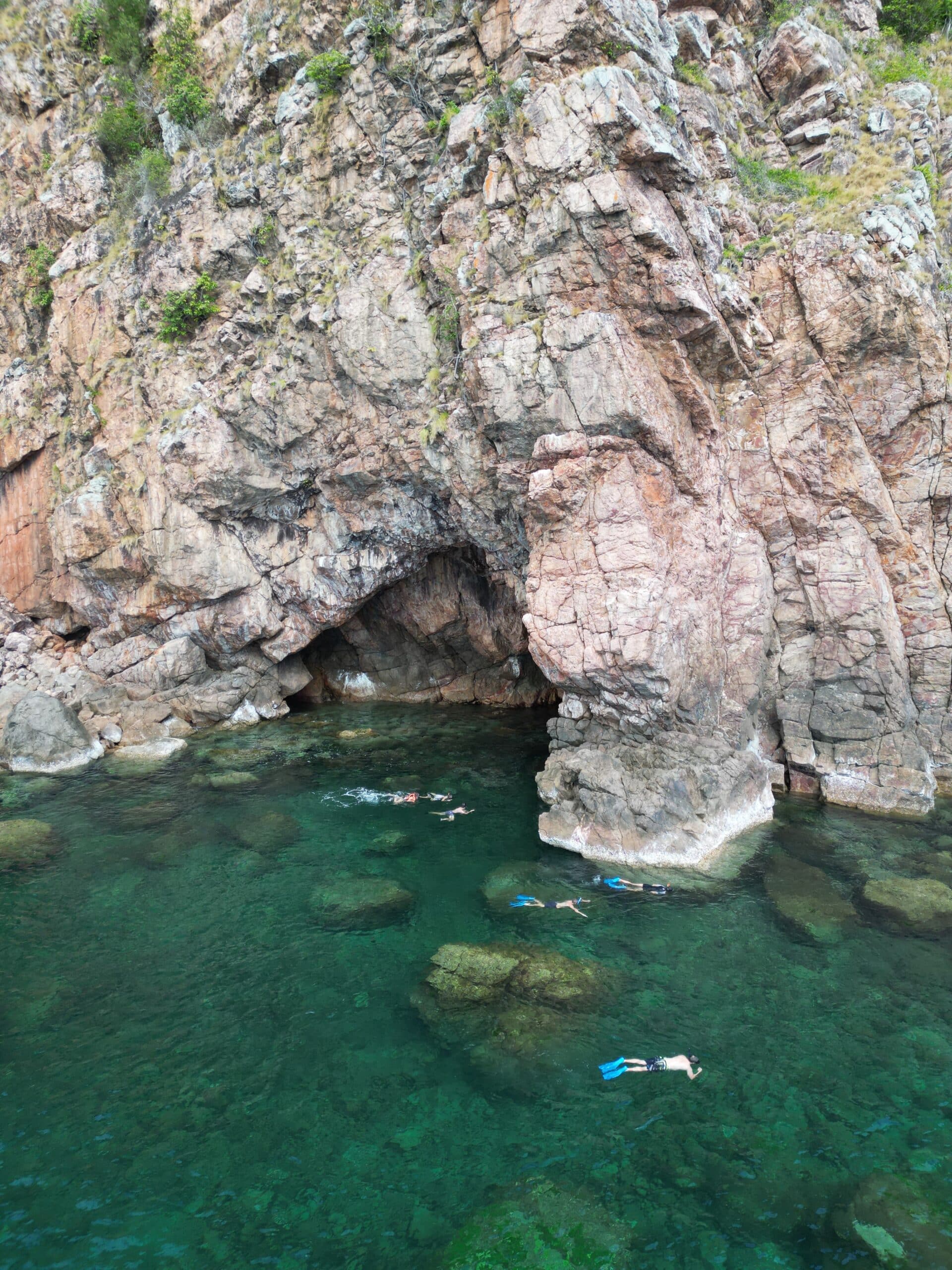Schnorchler schwimmen in der Nähe einer großen Felsklippe mit einer Höhlenöffnung. Das Wasser ist klar und gibt den Blick auf unter Wasser liegende Felsen frei. Die Felswand ist schroff und steil und an der Spitze mit spärlicher Vegetation bedeckt. Diese faszinierende Szenerie könnte leicht ein Höhepunkt jeder Segelreise nach Griechenland sein und strahlt Abenteuer und natürliche Schönheit aus.