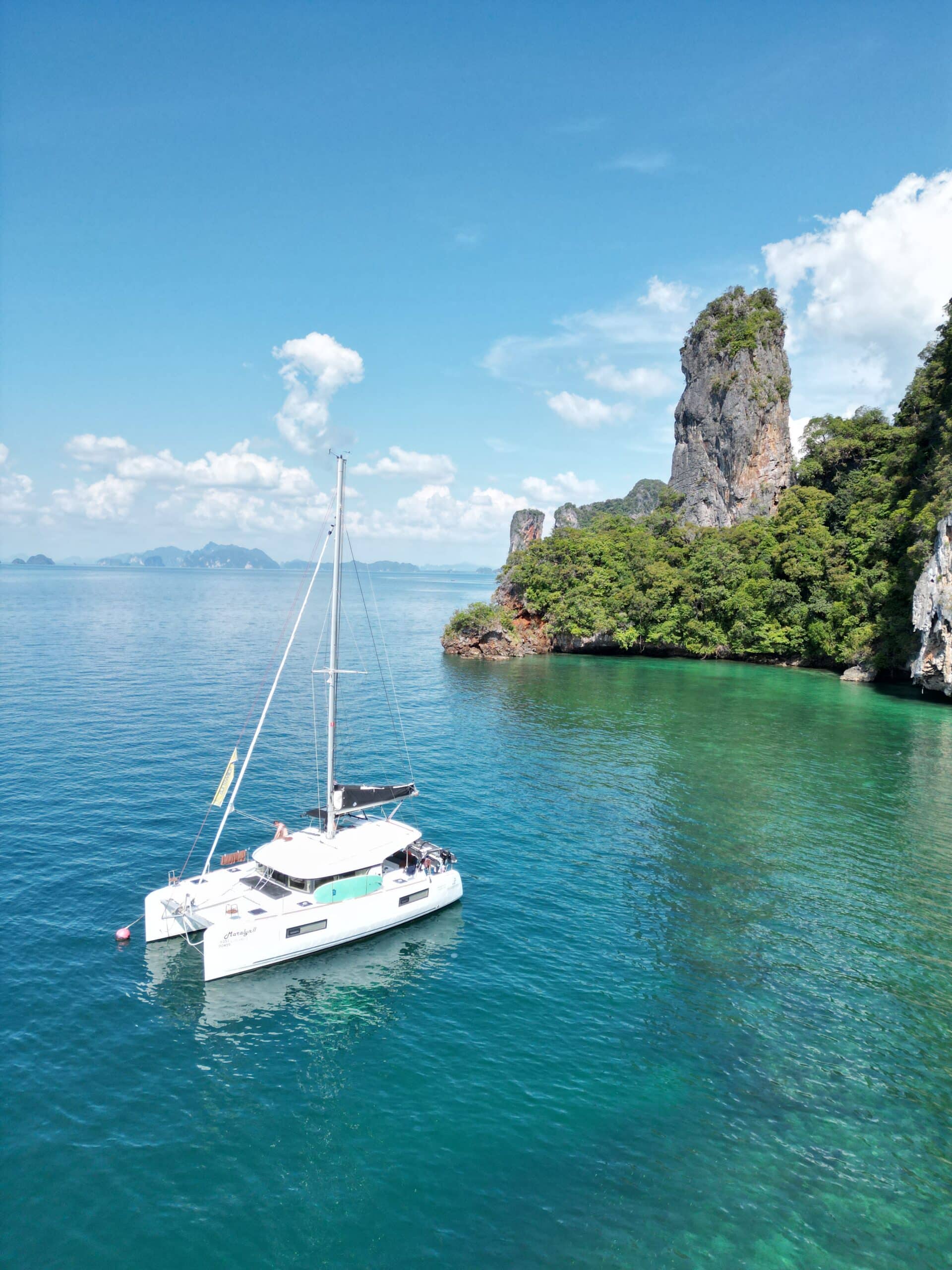 Ein weißer Katamaran segelt auf klarem, türkisfarbenem Wasser in der Nähe einer üppigen, grünen Insel mit hoch aufragenden Felsformationen unter einem strahlend blauen Himmel. Diese Segelreise Griechenland bietet eine heitere, malerische Umgebung mit ruhigem Meer und malerischer Kulisse.