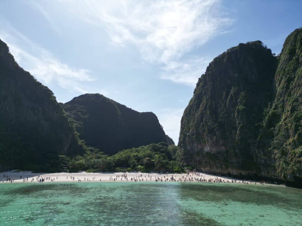 Ein malerischer Strand liegt eingebettet zwischen hoch aufragenden, üppig grünen Klippen unter einem strahlend blauen Himmel. Das klare, türkisfarbene Wasser erstreckt sich im Vordergrund, während sich eine Vielzahl von Menschen am Sandstrand versammelt hat und die malerische Umgebung genießt, die an eine Segelreise nach Griechenland erinnert.