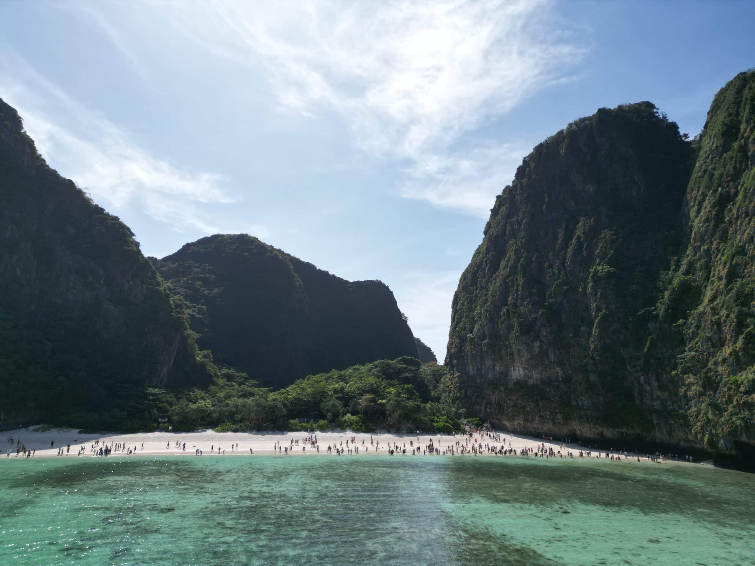 Eine wunderschöne tropische Strandszene mit klarem türkisfarbenem Wasser im Vordergrund. Der Strand ist voller Menschen und wird auf beiden Seiten von üppigen, grünen Klippen gesäumt, was an ein Segelreise-Abenteuer in Griechenland unter einem strahlend blauen Himmel mit dünnen Wolken erinnert.