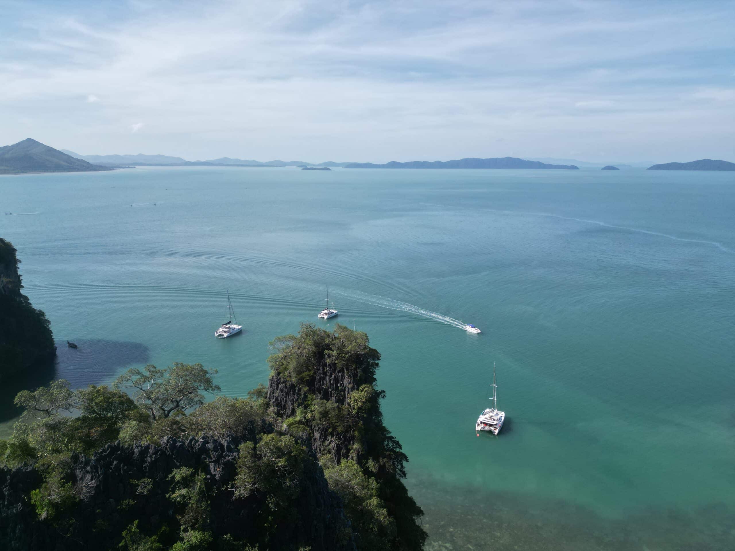 Luftaufnahme eines ruhigen türkisfarbenen Meeres mit vier Segelbooten in der Nähe felsiger, baumbedeckter Inseln, perfekt für eine Segelreise nach Griechenland. Der Himmel ist klar mit einigen Wolken und am Horizont sind in der Ferne Berge zu sehen.