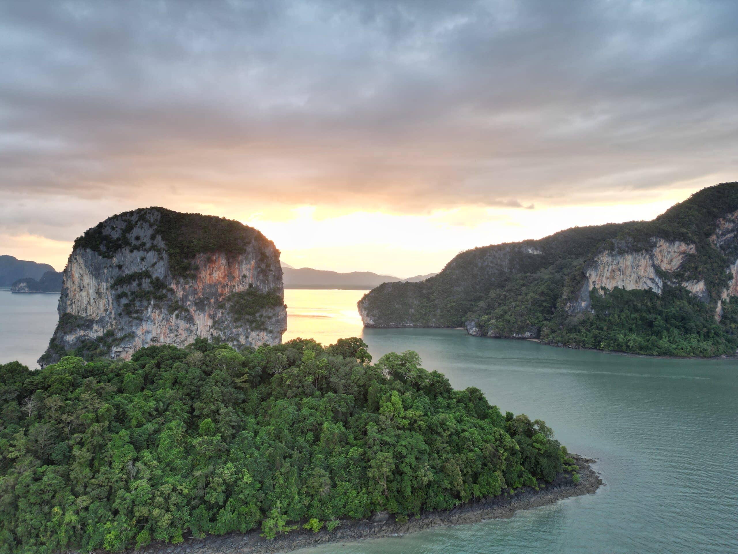 Luftaufnahme einer malerischen tropischen Bucht bei Sonnenuntergang mit hoch aufragenden Felsformationen, die von üppiger grüner Vegetation bedeckt sind. Die Sonne geht hinter den Klippen unter und wirft einen warmen Schein auf das ruhige Wasser und die bewaldeten Inseln darunter, was an eine ruhige Segelreise nach Griechenland unter einem teilweise bewölkten Himmel erinnert.