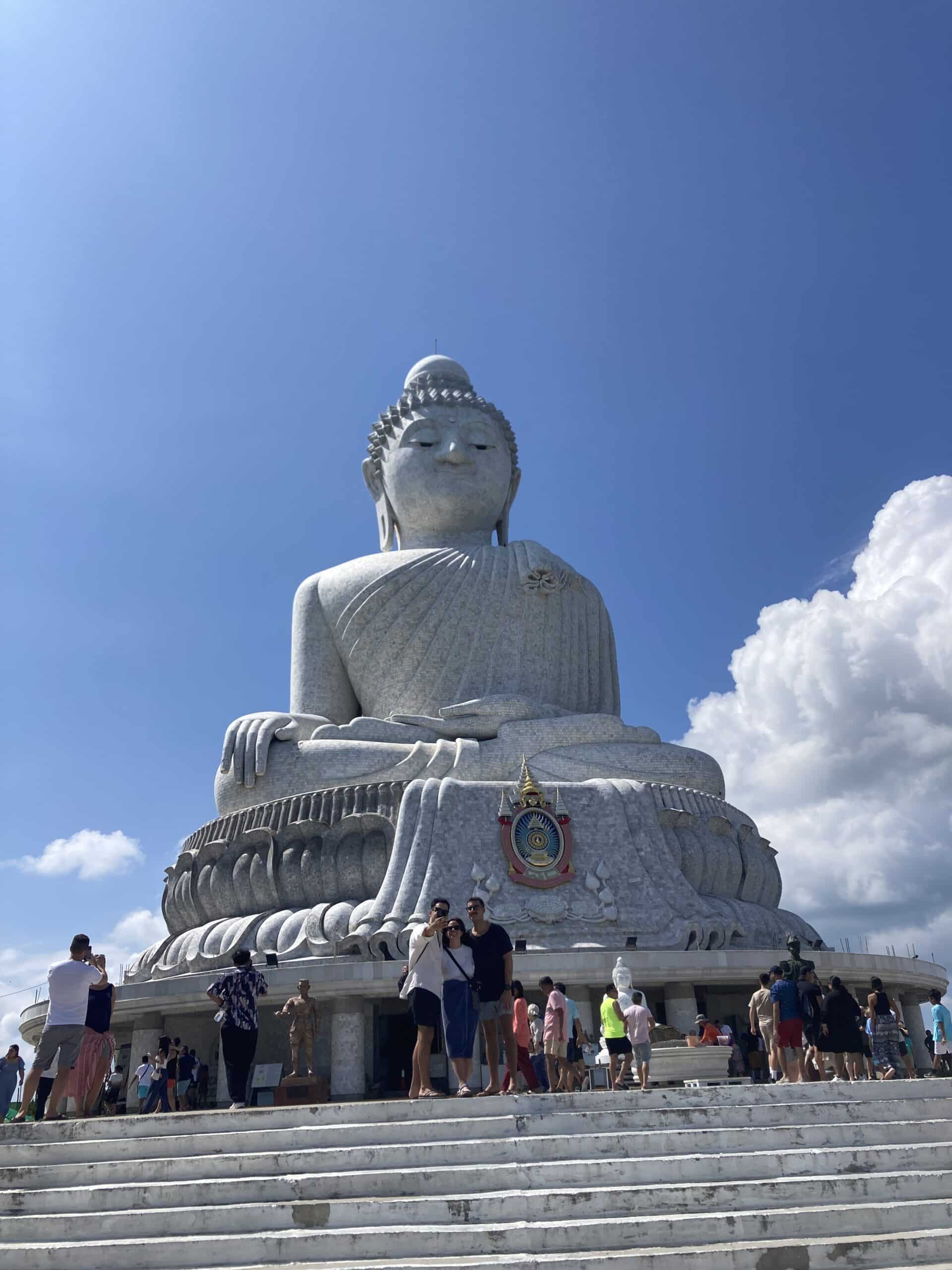 An einem klaren Tag versammeln sich Besucher am Fuß einer großen, sitzenden Buddhastatue aus Stein. Die Statue steht auf einer Plattform, zu der eine Treppe hinaufführt, unter einem strahlend blauen Himmel mit ein paar vereinzelten Wolken. Die Leute machen Fotos und laufen umher, was sie an ihre Erfahrungen bei der Segelreise nach Griechenland erinnert.