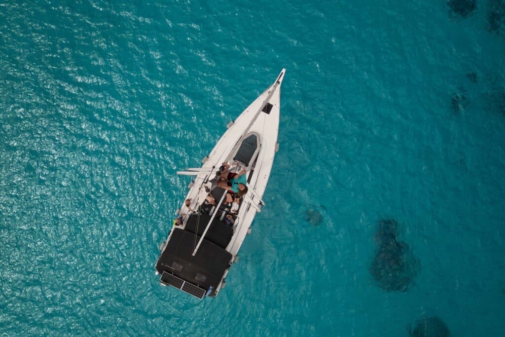 Luftaufnahme eines Segelboots mit einigen Personen an Deck, die während ihrer Segelreise Griechenland in klarem türkisfarbenem Wasser treiben. Der weiße Rumpf des Bootes kontrastiert mit dem leuchtend blauen Meer und schafft eine ruhige und malerische Szene. Mitsegeln.