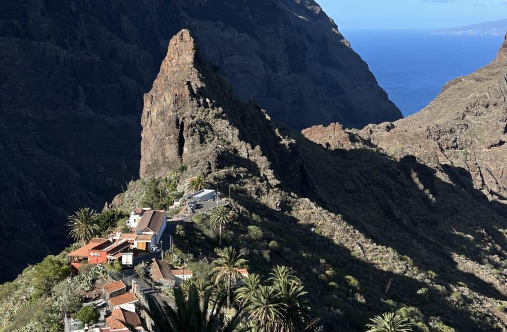 Zu sehen ist ein grüner Bergzipfel in der Nähe von der Küste. Umrundet von weiteren Bergen und kleinen Häusern und Palmen. Der Ort heißt Masca. Masca ist eines der malerischsten Dörfer Teneriffas und bietet atemberaubende Ausblicke auf die umliegenden Berge und Täler. Die steilen Klippen und das grüne Tal machen diesen Ort zu einem perfekten Fotomotiv. Die traditionelle kanarische Architektur und die spektakuläre Landschaft schaffen eine einzigartige Kulisse für Deine Fotos.