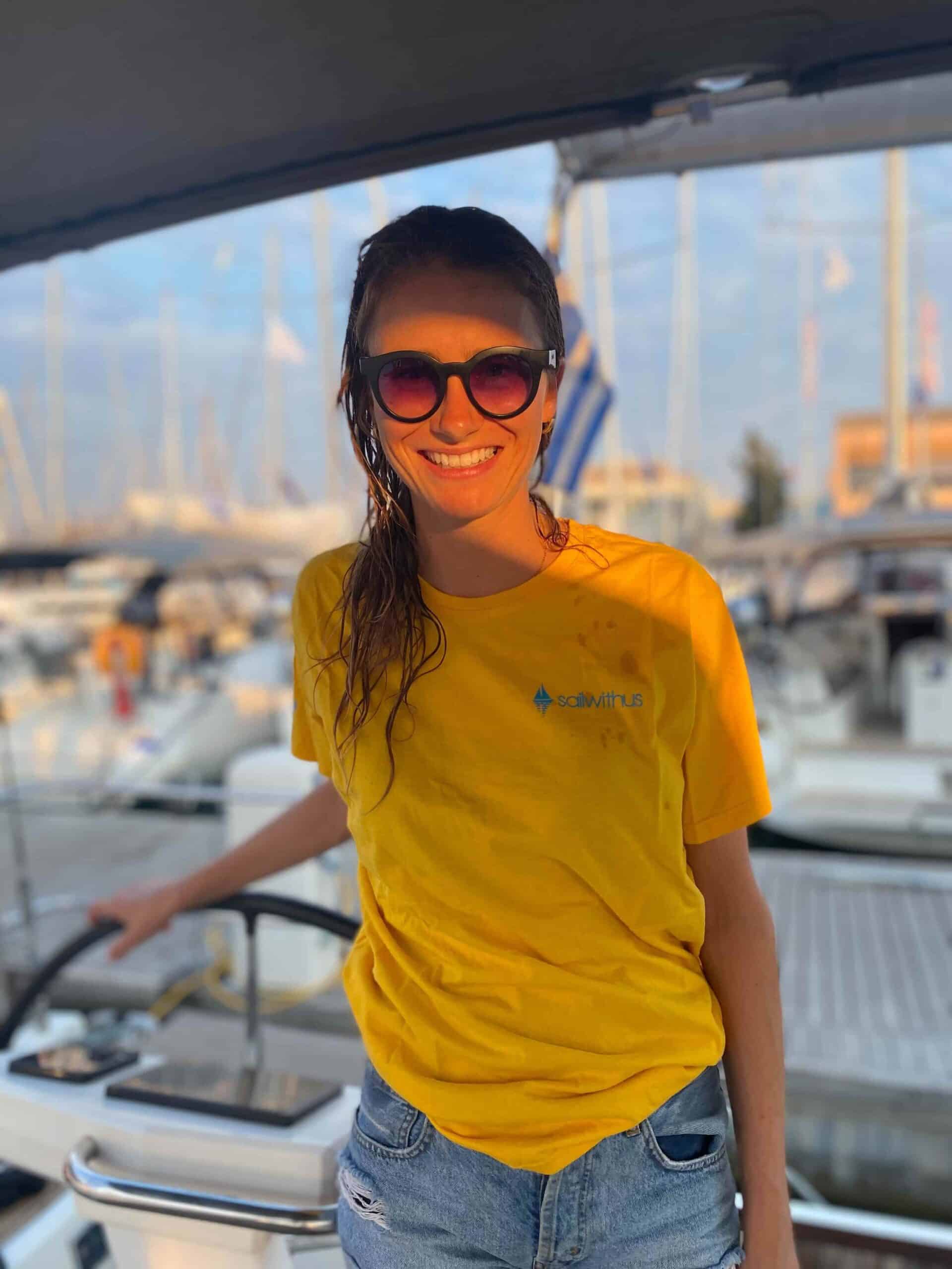 Eine Frau mit Sonnenbrille, gelbem T-Shirt und Jeansshorts steht lächelnd am Steuer eines Bootes während ihres Segelurlaubs. Im Hintergrund sind mehrere angedockte Boote und ein Yachthafen zu sehen. Das Sonnenlicht sorgt für eine warme Atmosphäre.