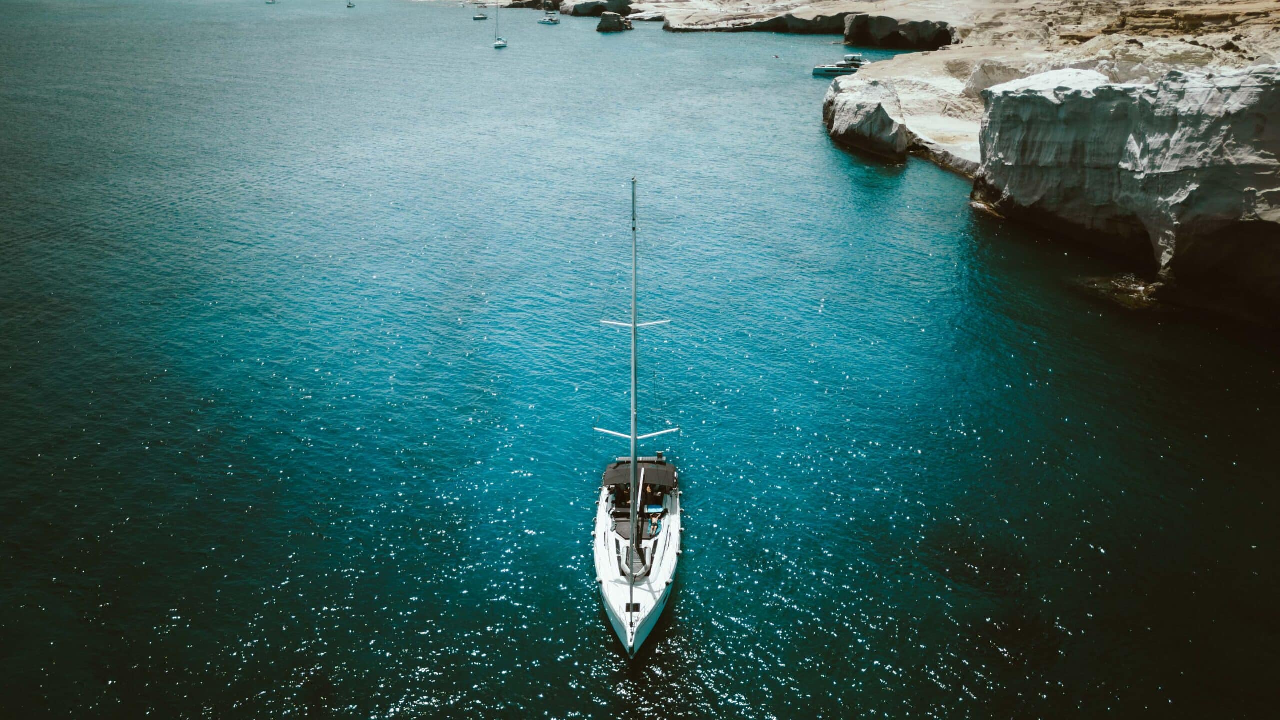 Ein weißes Segelboot schwimmt auf einem klaren, tiefblauen Meer nahe einer felsigen Küste mit weißen Klippen. Das Wasser glitzert im hellen Sonnenlicht und in der Ferne sind andere kleinere Boote zu sehen. Es ist eine ideale Kulisse für Skippertraining im Mittelmeer und macht die Szenerie sowohl ruhig als auch malerisch.