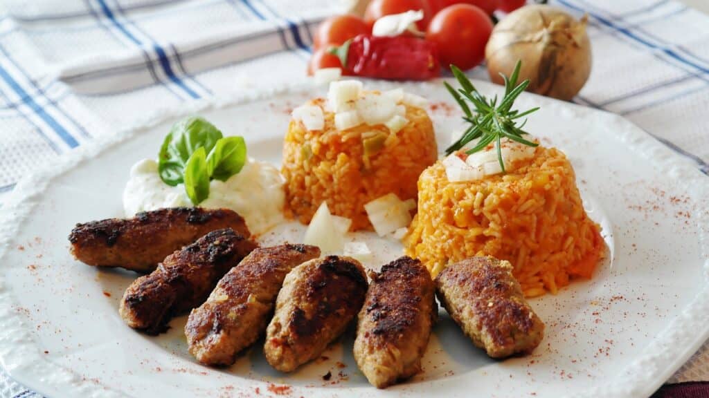 Ćevapčići sind würzigen Hackfleischröllchen aus Rind oder Lamm werden traditionell gegrillt und mit Zwiebeln und Ajvar, einer scharfen Paprikasauce, serviert. Dabei ist roter reis. alles ist schön auf einem Teller angerichtet. im Hintergrund sieht man Tomaten und Zwiebeln.