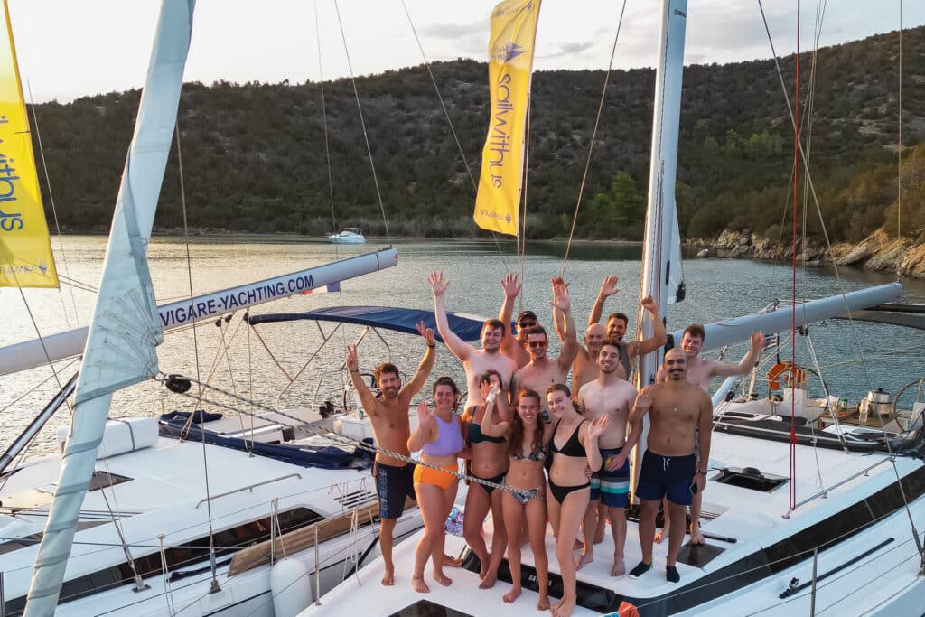 Eine Gruppe von Menschen steht lächelnd und winkend auf dem Deck einer weißen Yacht. Sie tragen Badebekleidung und scheinen einen sonnigen Tag während ihres Segelurlaubs auf Sardinien zu genießen. Neben ihnen ist eine weitere Yacht zu sehen, im Hintergrund sind malerische Hügel und Bäume zu sehen.