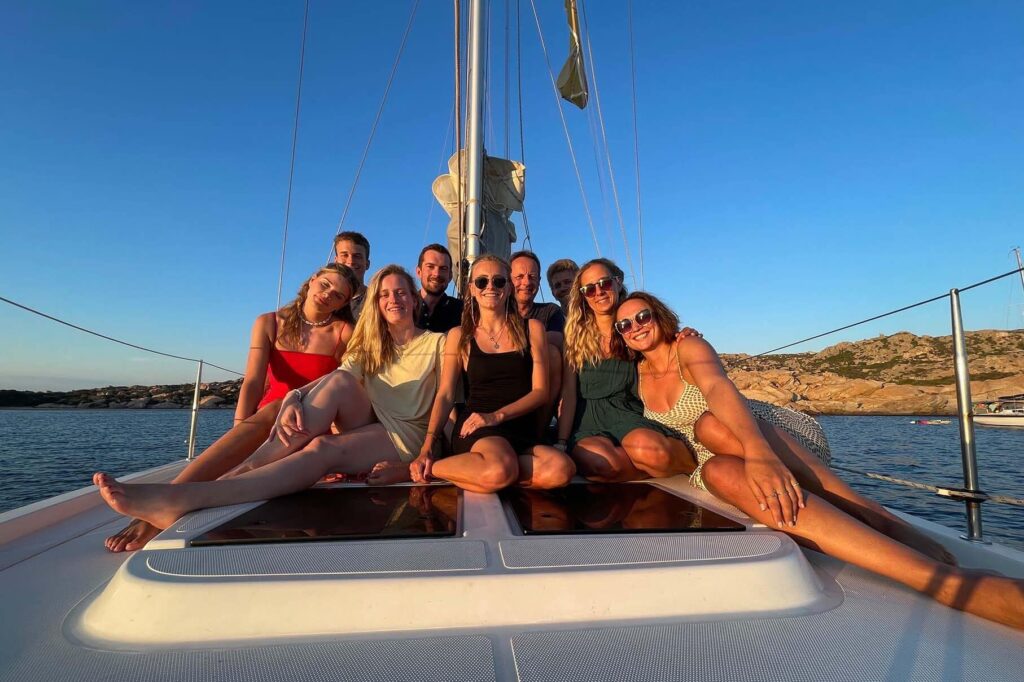 Eine Gruppe von acht Freunden posiert während ihrer Segelreise bei Sonnenuntergang auf dem Vorderdeck eines Segelboots und lächelt freudig vor der malerischen Kulisse des ruhigen Meeres und der felsigen Küste.