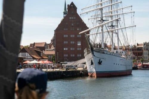 Eine Person beobachtet an einem sonnigen Tag ein großes weißes Segelschiff, das in einem Hafen mit traditionellen roten Backsteingebäuden und einem Kirchturm im Hintergrund vor Anker liegt. Kulinarische Höhepunkte bei Hafenstopps an der Ostsee