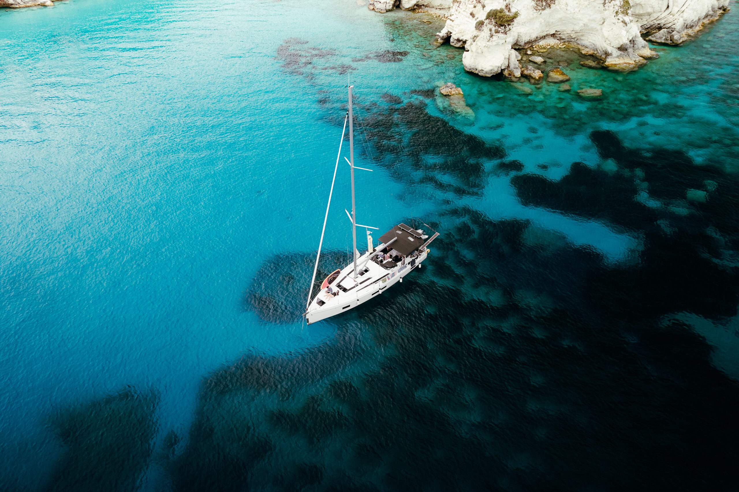 Ein Segelboot ankert in einer ruhigen, türkisfarbenen Bucht, umgeben von teilweise unter Wasser liegenden Felsen und weißen Klippen – perfekt für eine ruhige Pause während einer Atlantiküberquerung. Das Wasser variiert in Blautönen, mit Flecken von dunklerem Blau und Grün um das Boot herum. Kombinationsreise und Segelreise.