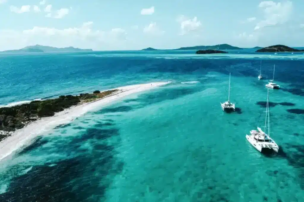Luftaufnahme eines schmalen Sandstrands mit kristallklarem, türkisfarbenem Wasser, umgeben von mehreren Segelyachten vor einer üppig grünen Insel. Karibik Segeltörn in der Nebensaison