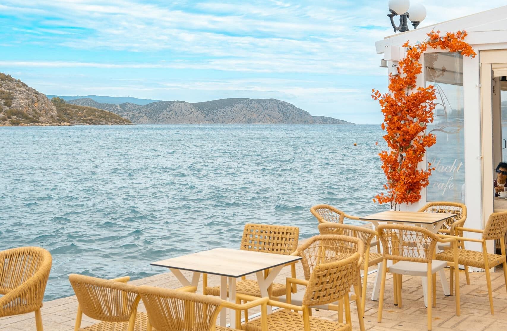 Sunny seaside café terrace in Ermioni, Greece, with wicker chairs and vibrant decor.
