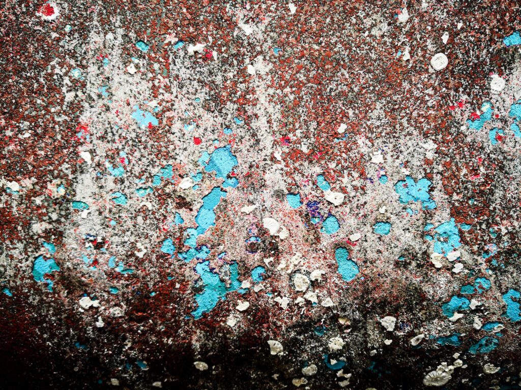 Eine Nahaufnahme einer strukturierten Oberfläche mit rostbraunem Hintergrund, die an ein abstraktes Kunstwerk erinnert, das vom Mistralwind geformt wurde. Es ist mit verschiedenen Farben gesprenkelt, darunter weiße Flecken, leuchtendes Blau und ein Hauch von Rot und Grün. Die Oberfläche wirkt abgenutzt und verwittert.