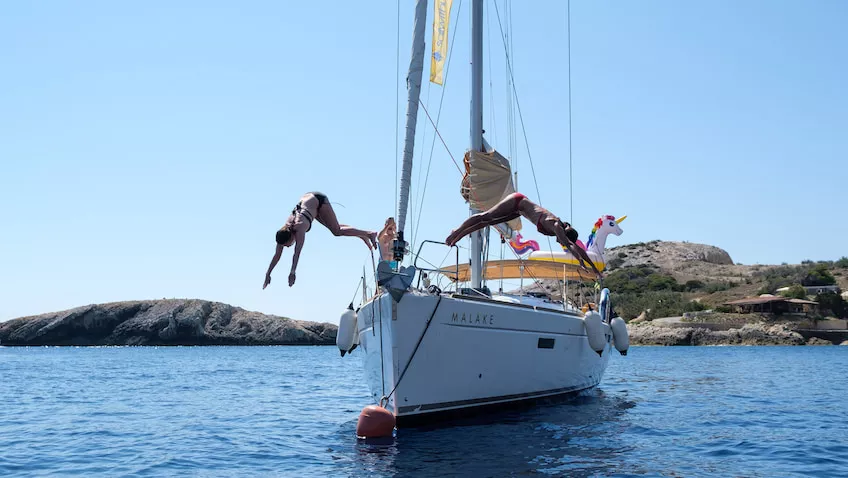 Zwei Personen springen von einer Segelyacht ins Meer, während eine andere Person vom Boot aus zusieht, vor der Kulisse eines klaren blauen Himmels und einer felsigen Küste.