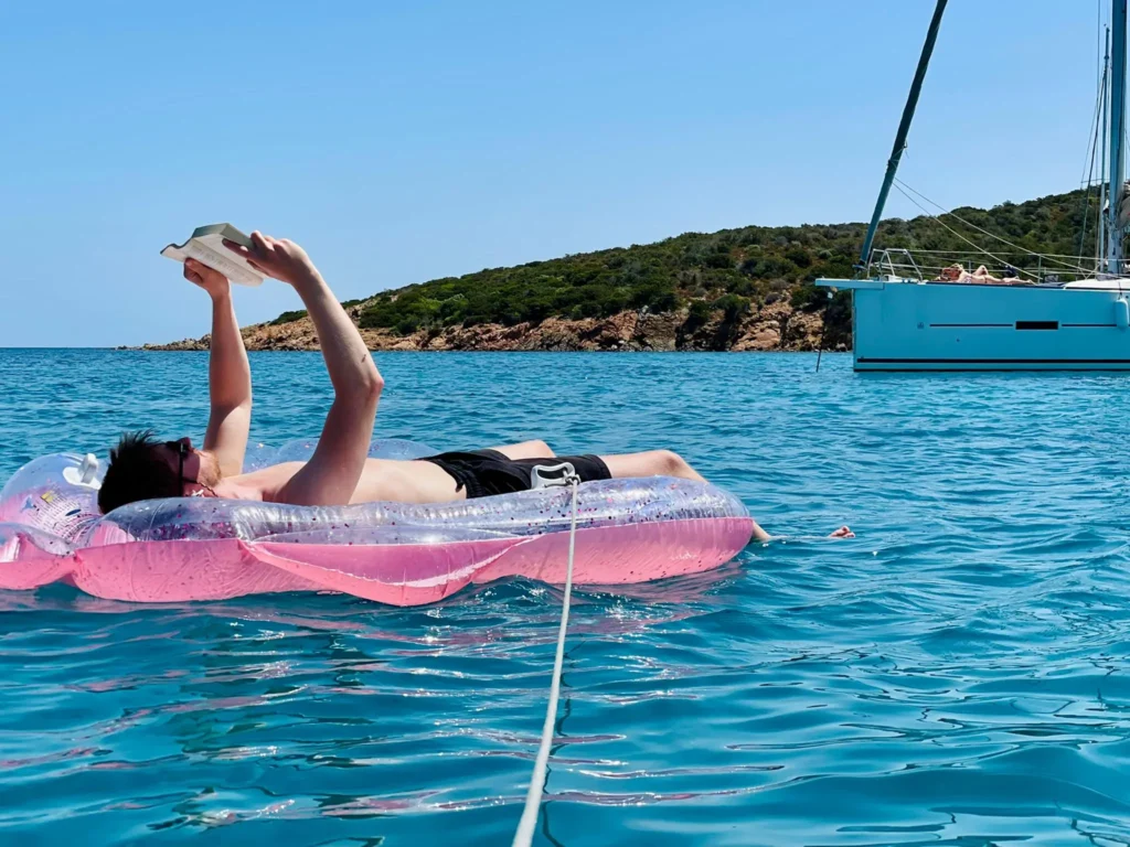 Eine Person liegt während ihres Segelurlaubs auf einem rosa aufblasbaren Ring im klaren, blauen Meereswasser und macht ein Selfie mit einem Smartphone. In der Nähe ist unter einem klaren Himmel ein Segelboot vertäut.
