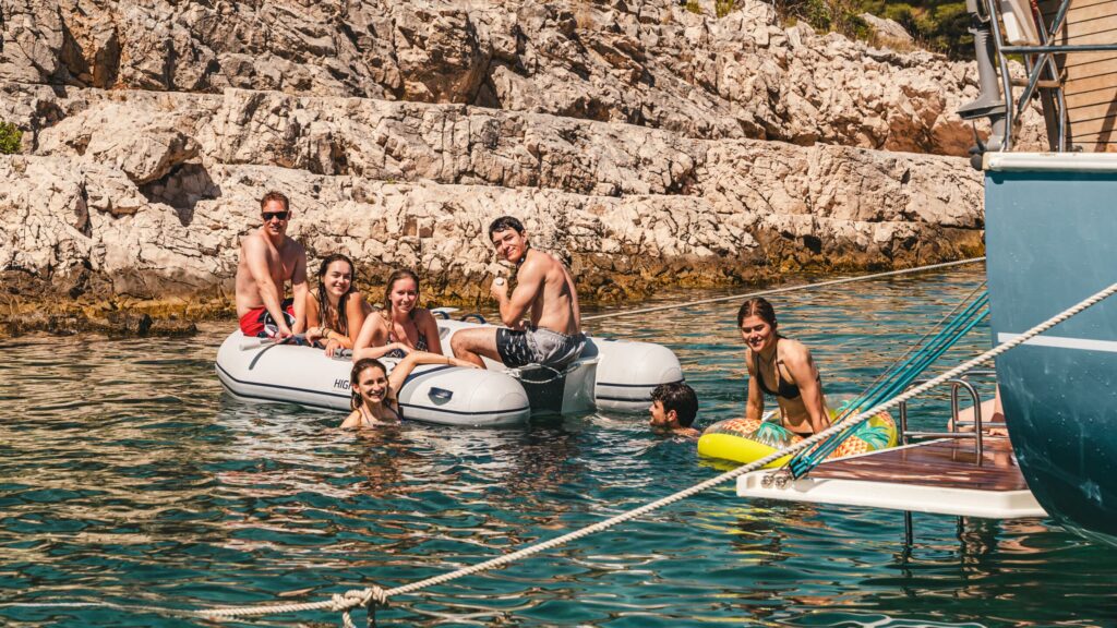 Eine Gruppe von Menschen genießt einen sonnigen Tag im klaren blauen Wasser nahe einer felsigen Küste. Einige schwimmen, während andere bunte Schwimmflügel und Poolnudeln benutzen. Bäume und Sträucher sind auf dem felsigen Hintergrund zu sehen, was an Mitsegeln in Kroatien erinnert. Die Szene ist lebhaft und entspannt.
