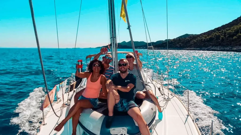 Vier Freunde genießen einen sonnigen Tag auf einer Segelyacht und winken in die Kamera, während sie vom klaren blauen Himmel und dem glitzernden Meerwasser umgeben sind.