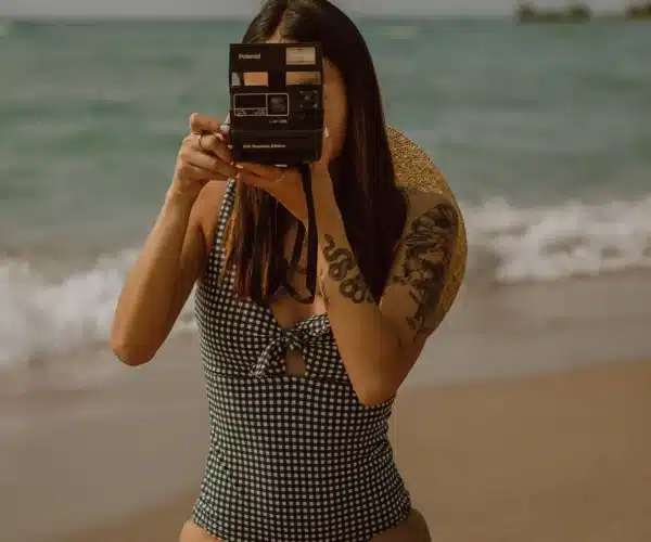 Eine Frau in einem karierten Kleid steht am Strand und hält eine alte Polaroid-Kamera vor ihr Gesicht, bereit, ein Foto zu machen. Ihr tätowierter Arm ist sichtbar, als sie sich darauf vorbereitet,