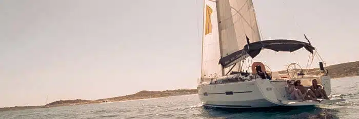 Eine Segelyacht mit gelbem Segel kreuzt auf ruhigen Gewässern, während mehrere Passagiere auf dem Deck sitzen und das sonnige Wetter genießen.