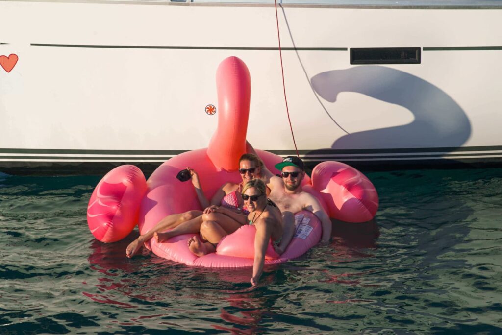 Drei Personen entspannen auf einem großen rosa Flamingo-Schwimmreifen neben einem Boot im Wasser und genießen die Sonne. Sie scheinen in fröhlicher Stimmung zu sein, während sie die ruhige Meereskulisse während ihrer