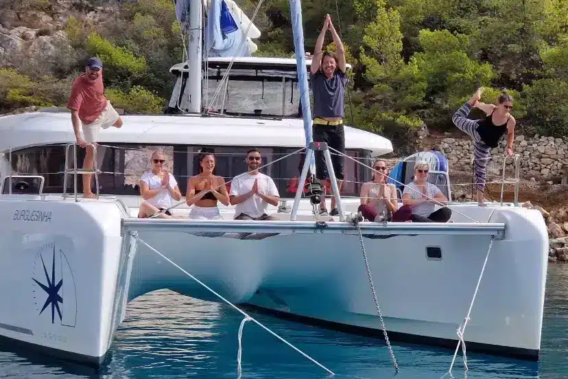 Eine Gruppe von Menschen vergnügt sich auf einer Segelyacht, die in der Nähe einer felsigen Küste vor Anker liegt. Einige sitzen, andere stehen oder posieren, alle wirken fröhlich unter dem sonnigen Himmel.