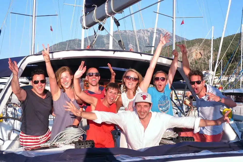Eine fröhliche Gruppe von acht Freunden posiert auf einer Segelyacht, mit feierlich erhobenen Armen und einem Lächeln vor der malerischen Kulisse von Bergen und klarem Himmel.