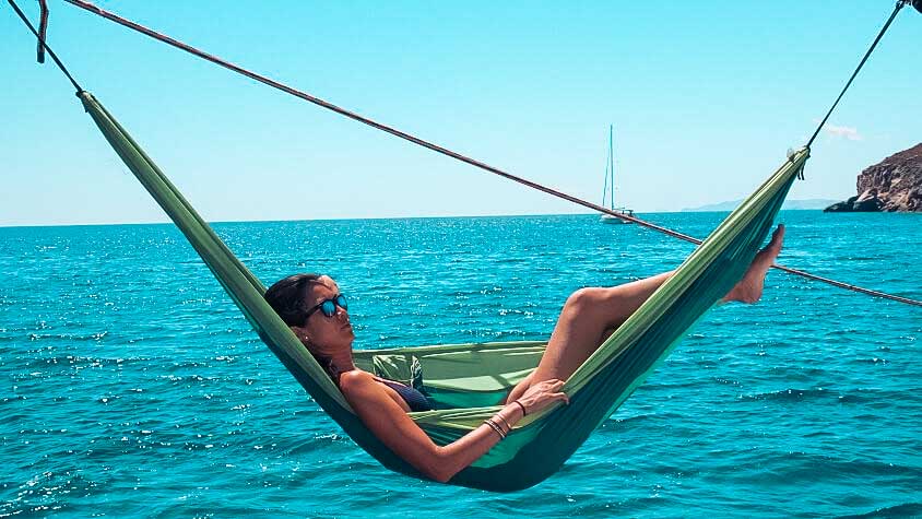 Eine Person entspannt sich in einer grünen Hängematte über dem klaren, blauen Meer unter einem strahlend sonnigen Himmel. In der Ferne sind Hügel und im Hintergrund eine Segelyacht zu sehen.