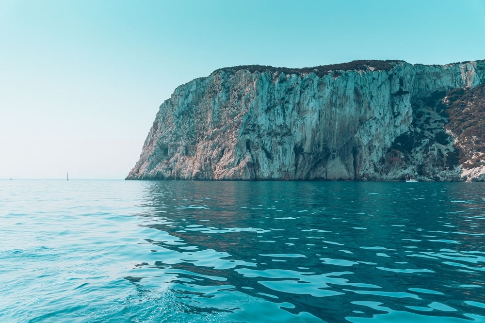 Türkisfarbenes Wasser mit sanften Kräuselungen vor einer hoch aufragenden Klippe unter klarem Himmel, das bei einer Segelreise Schatten und Lichteffekte auf die Felswand wirft.