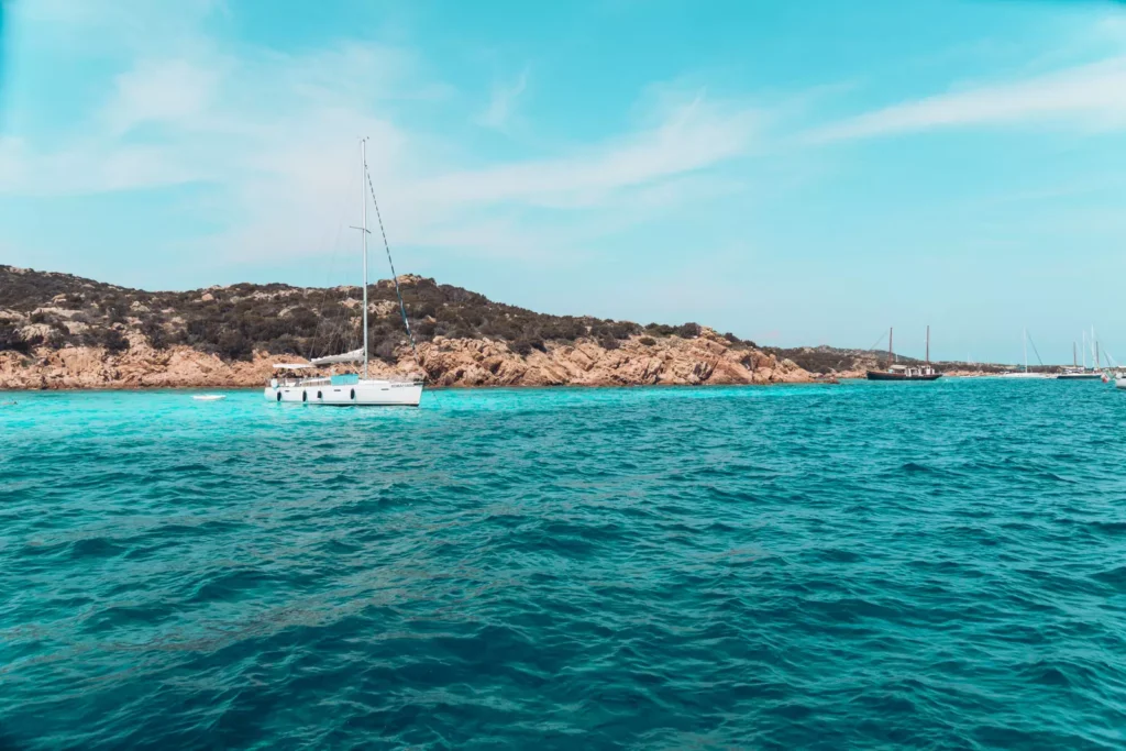 Eine ruhige Meereslandschaft mit einer weißen Segelyacht, die unter einem klaren blauen Himmel in der Nähe einer felsigen Küste vor Anker liegt. In der Ferne sind weitere Segelboote zu sehen.