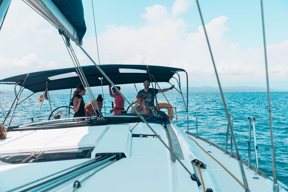 Vier Personen genießen einen sonnigen Tag auf einem Segelboot während eines Segeltörns, mit offenem Wasser und klarem Himmel im Hintergrund. Der Blick geht vom Bug zum Heck. Seekrankheit Tipps