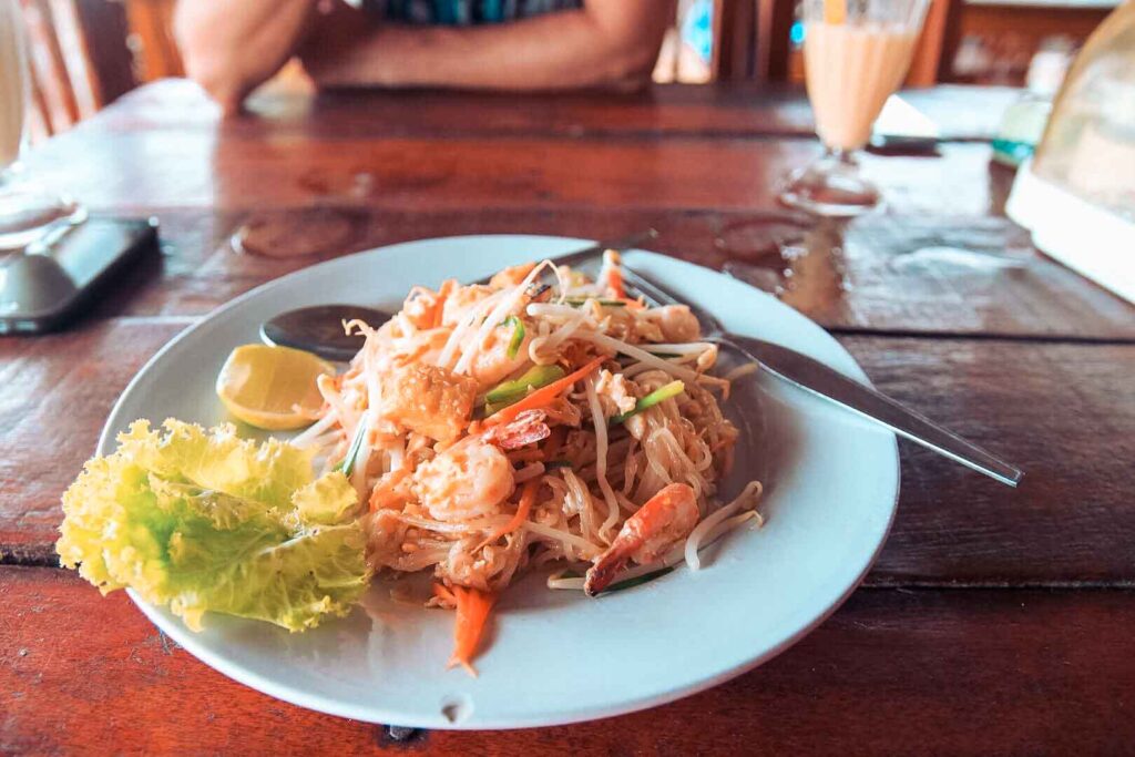 Ein Teller mit Garnelen-Pad-Thai, einer Limettenscheibe und Salat auf einem Holztisch, mit einem verschwommenen Hintergrund, der eine Person im Segelurlaub und ein Getränk zeigt.