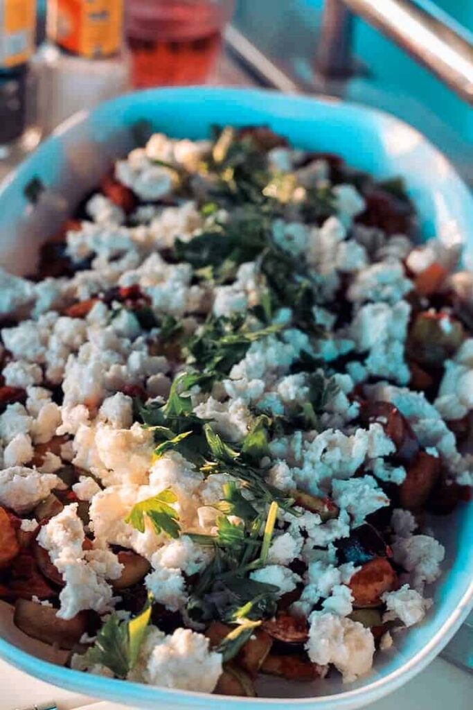 Ein Gericht aus Bratkartoffeln, garniert mit geriebenem Käse und frischen grünen Kräutern, serviert auf einer Küchentheke, die während einer Segelreise von natürlichem Sonnenlicht beleuchtet wird.