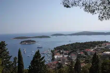 Eine malerische Aussicht von einem Hügel, die eine Küstenstadt mit Dächern, üppigem Grün und mehreren Booten, darunter Segelyachten, in einem ruhigen blauen Meer zeigt, mit Inseln im Hintergrund unter einem klaren Himmel