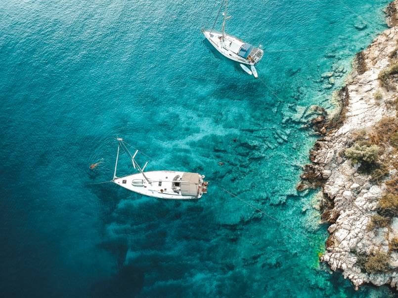 Luftaufnahme von zwei Segelyachten, die in der Nähe einer felsigen Küste mit klarem, türkisfarbenem Wasser vor Anker liegen.