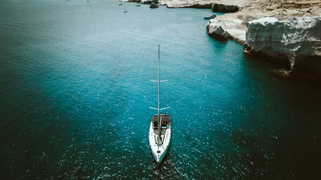 Ein weißes Segelboot schwimmt auf einem klaren, tiefblauen Meer nahe einer felsigen Küste mit weißen Klippen. Das Wasser glitzert im hellen Sonnenlicht und in der Ferne sind andere kleinere Boote zu sehen. Es ist eine ideale Kulisse für Skippertraining im Mittelmeer und macht die Szenerie sowohl ruhig als auch malerisch.